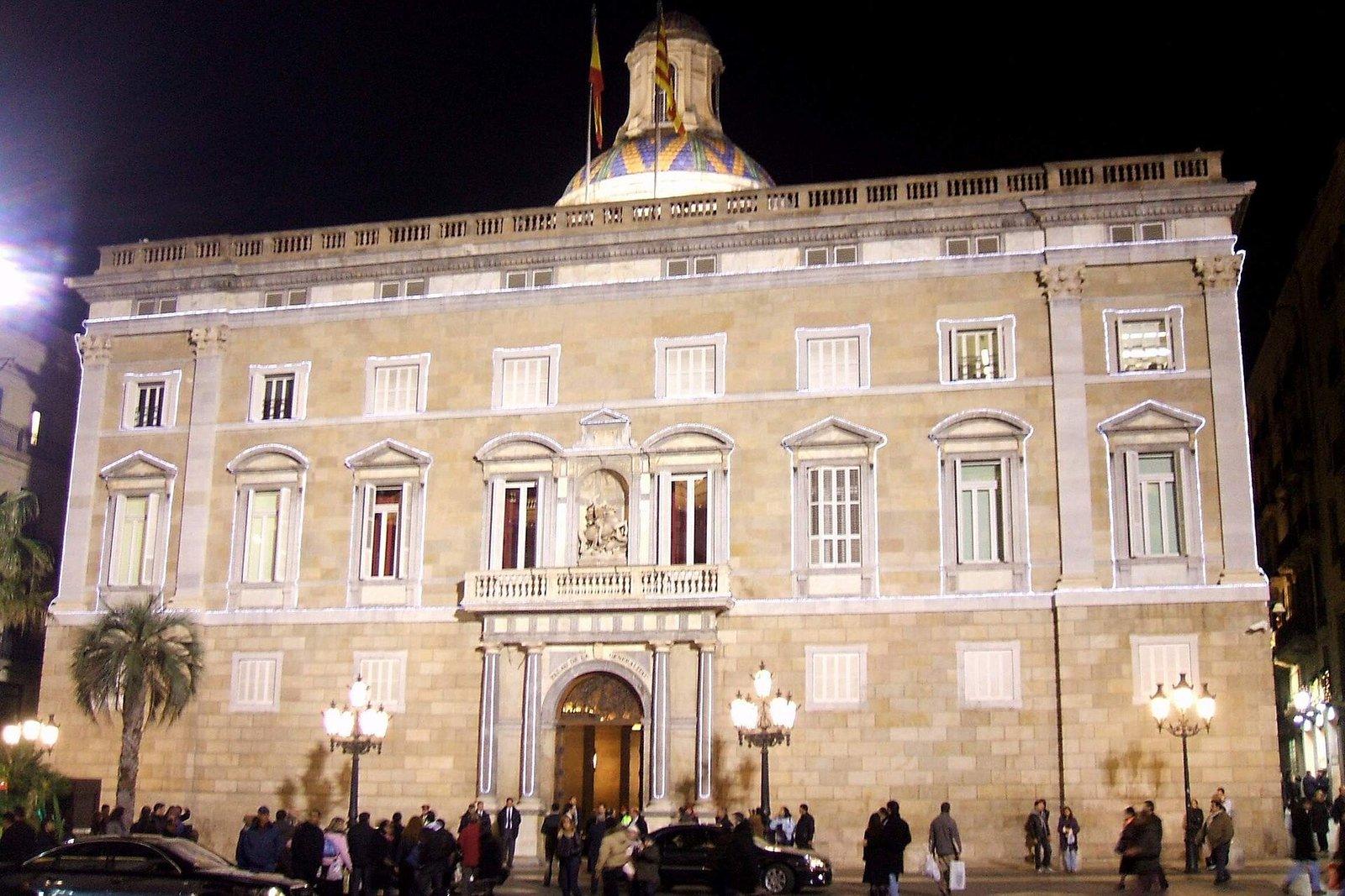 Fachada iluminada de noche del Palau de la Generalitat en Barcelona, con banderas, cúpula y gente en la plaza.