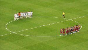 Dos equipos alineados junto al círculo central del campo antes de un partido, con el árbitro y el balón en el centro.