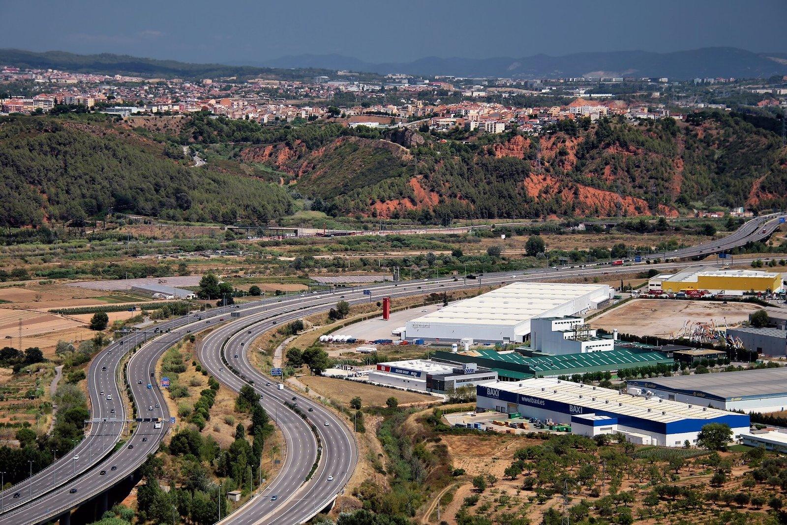 Vista aérea de la autovía A-2 y la autopista AP-7 con tráfico fluido, naves industriales y colinas al fondo.